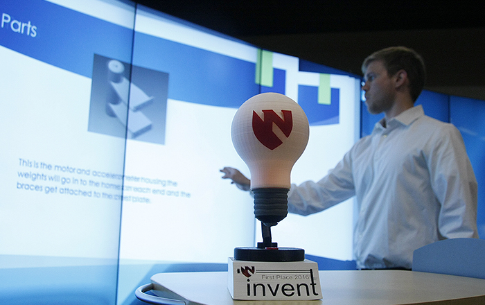 In the foreground is the 2016 Invent-a-Thon trophy, a 3D-printed model of a light bulb that is lit with LEDs. In the background, an officer of UNMC's 3D Maker's Club, Will Payne, prepares one of the competitors’ presentations on the iEXCEL center’s MultiTaction Wall.